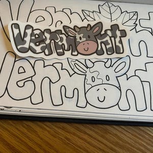 May include: A coloring book page with the word "Vermont" written in large, outlined letters. The word is repeated twice, with a cow sticker placed over the second word. The cow sticker has black and white spots and a pink nose.