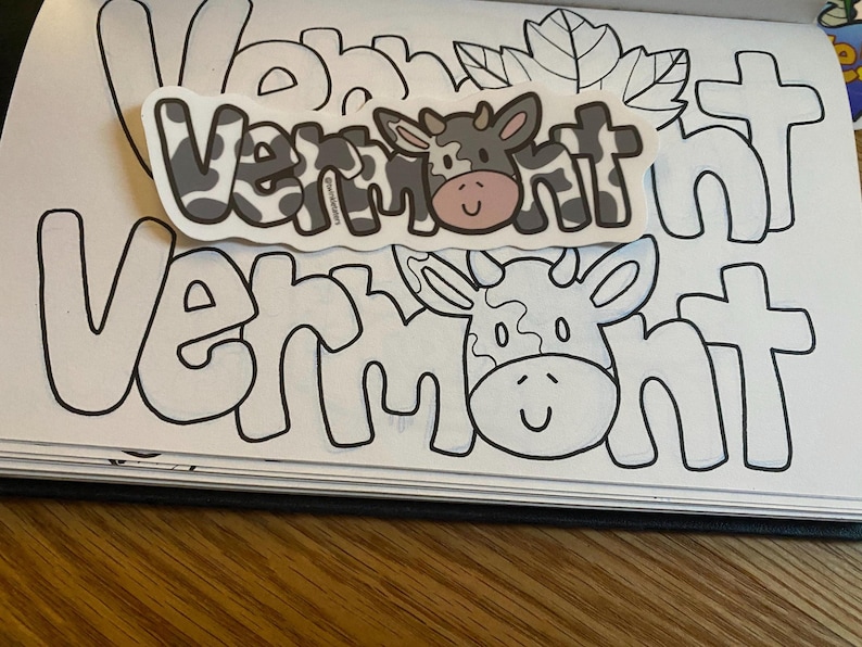 May include: A coloring book page with the word "Vermont" written in large, outlined letters. The word is repeated twice, with a cow sticker placed over the second word. The cow sticker has black and white spots and a pink nose.