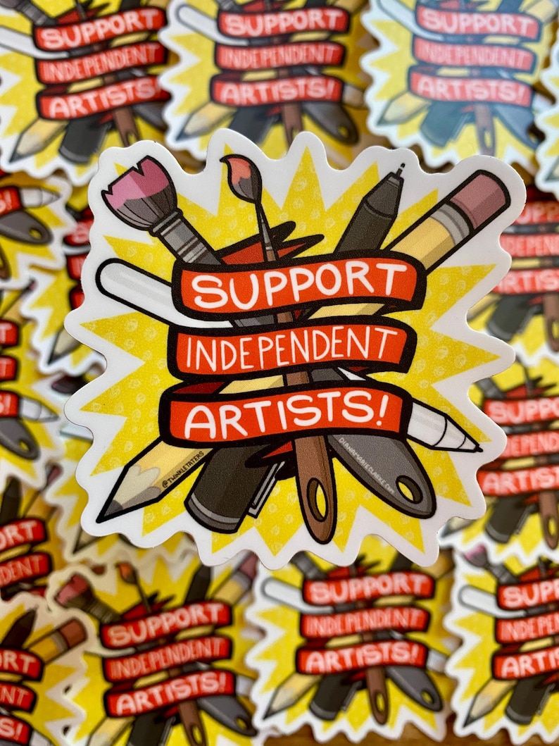 Puede incluir: Una pegatina con un dise&ntilde;o de estrella amarilla que presenta materiales de arte como pinceles, l&aacute;pices y bol&iacute;grafos. Una pancarta roja en el centro dice "SUPPORT INDEPENDENT ARTISTS!" en texto blanco. La pegatina es un llamado a apoyar a los artistas.