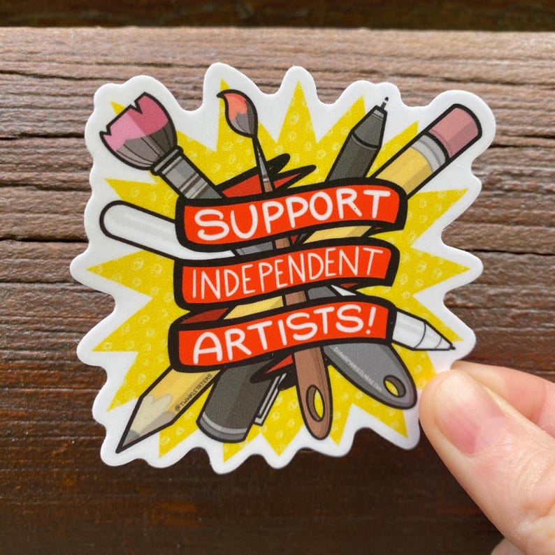 Puede incluir: Una pegatina con un dise&ntilde;o de explosi&oacute;n amarilla que presenta &uacute;tiles de arte como un pincel, un l&aacute;piz y un bol&iacute;grafo. Una pancarta roja en el centro dice "SUPPORT INDEPENDENT ARTISTS!" en texto blanco. La pegatina est&aacute; sujeta por una mano.