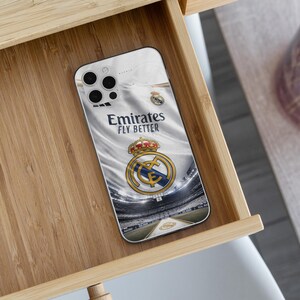 May include: A smartphone case featuring a white background with the Real Madrid football club crest and the text Emirates FLY BETTER. The design includes a graphic of a stadium interior, showcasing the team logo with a gold crown and blue details.