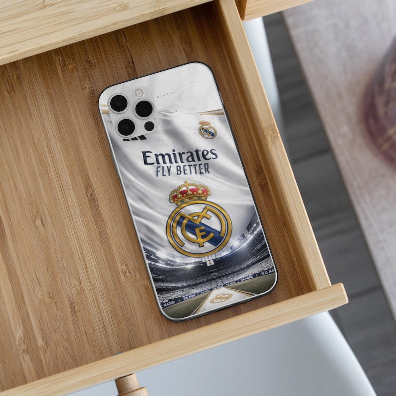 May include: A smartphone case featuring a white background with the Real Madrid football club crest and the text Emirates FLY BETTER. The design includes a graphic of a stadium interior, showcasing the team logo with a gold crown and blue details.