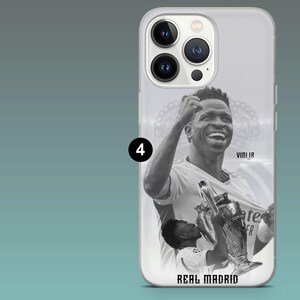 May include: A clear smartphone case featuring a black and white graphic of a soccer player celebrating with a trophy. The design includes the text VINI JR and REAL MADRID. The case has a cutout for a triple-lens camera system on the back.