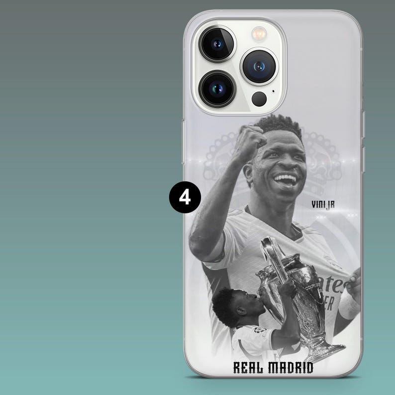 May include: A clear smartphone case featuring a black and white graphic of a soccer player celebrating with a trophy. The design includes the text VINI JR and REAL MADRID. The case has a cutout for a triple-lens camera system on the back.