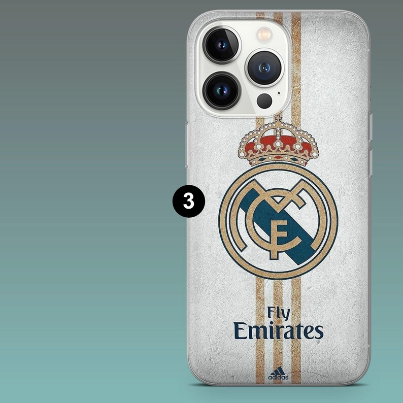 May include: A white smartphone case featuring the Real Madrid crest in gold and blue, topped with a red and gold crown. Three vertical gold stripes run down the center. The text Fly Emirates and a small Adidas logo are printed in dark blue.