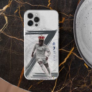 May include: A clear phone case featuring a graphic of a soccer player in a white jersey wearing a red crown. The design includes a large dark gray number seven, the text VINI JR, and the Real Madrid logo on a white background with abstract brush strokes.