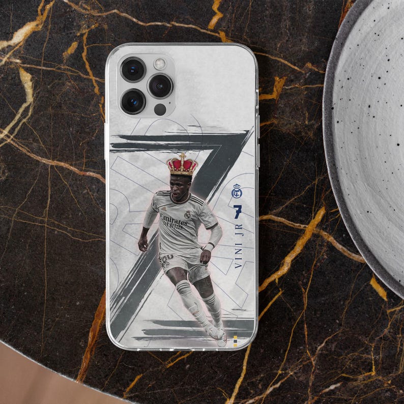 May include: A clear phone case featuring a graphic of a soccer player in a white jersey wearing a red crown. The design includes a large dark gray number seven, the text VINI JR, and the Real Madrid logo on a white background with abstract brush strokes.
