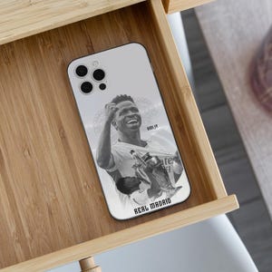 May include: A white smartphone case featuring a black and white graphic of a soccer player celebrating with a trophy. The text VINI JR is printed near the top right, and the text REAL MADRID is printed in bold black letters at the bottom.