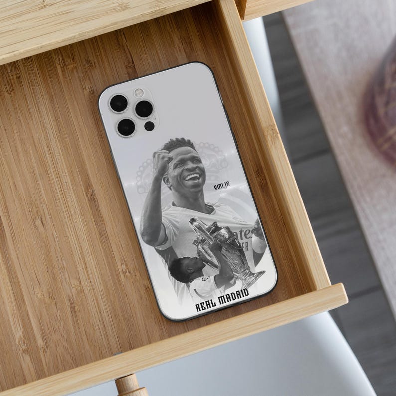 May include: A white smartphone case featuring a black and white graphic of a soccer player celebrating with a trophy. The text VINI JR is printed near the top right, and the text REAL MADRID is printed in bold black letters at the bottom.