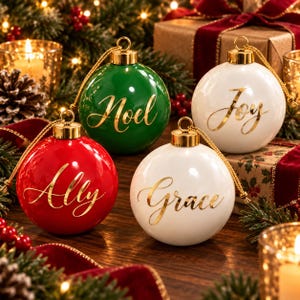 May include: Four round Christmas ornaments with gold caps and hanging strings. One green ornament features the gold text Noel, one red ornament features the gold text Ally, and two white ornaments feature the gold text Joy and Grace respectively.