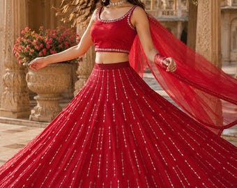 May include: A vibrant red lehenga set featuring a sleeveless crop top and a voluminous floor-length skirt. Both pieces are adorned with vertical rows of silver metallic embroidery and small mirror accents. A matching sheer red dupatta is draped over one arm.