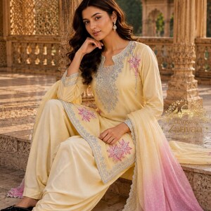 May include: A pale yellow salwar kameez set featuring intricate light blue embroidery around the neckline and cuffs, accented with pink floral motifs on the tunic and a matching dupatta with a pink ombre gradient and scalloped silver trim edges.