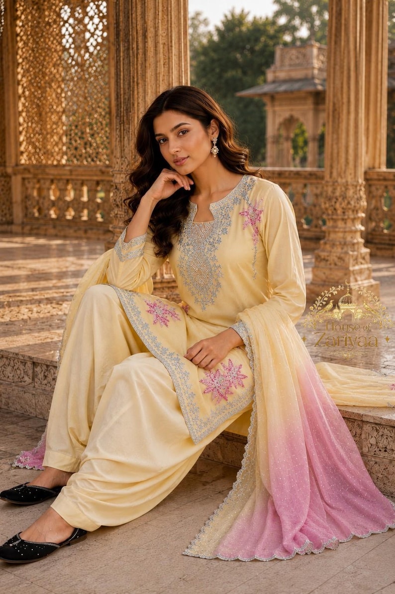 May include: A pale yellow salwar kameez set featuring intricate light blue embroidery around the neckline and cuffs, accented with pink floral motifs on the tunic and a matching dupatta with a pink ombre gradient and scalloped silver trim edges.