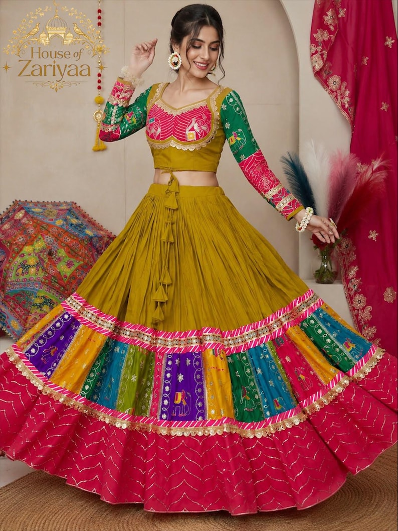May include: A mustard yellow lehenga choli set featuring a long, pleated skirt with a multi-colored patchwork hem in purple, blue, green, and yellow, finished with a wide magenta border. The long-sleeved blouse has pink and green patterned accents.