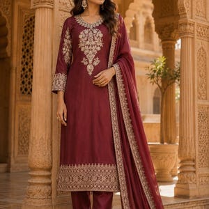 May include: A deep maroon salwar kameez set featuring intricate gold embroidery on the neckline, sleeves, and hem. The ensemble includes a matching maroon dupatta with a gold embroidered border and coordinating straight-leg trousers with gold detailing.