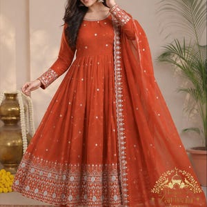 May include: A rust-orange Anarkali dress featuring long sleeves with intricate silver embroidery at the cuffs, a round neckline, and a flared skirt adorned with small mirror accents and a wide, detailed silver embroidered border at the hem.