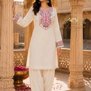 May include: An off-white long-sleeved tunic and matching salwar pants set. The tunic features pink and green floral embroidery around the neckline and on the sleeve cuffs. The pants have a gathered, pleated design with pink trim at the ankles.