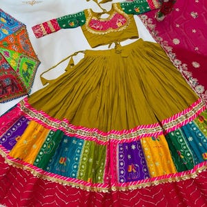 May include: A mustard yellow lehenga choli set featuring a pleated skirt with a multi-colored patchwork border of elephant motifs in green, purple, and yellow, accented with gold trim. The blouse has green sleeves and a pink embroidered neckline.