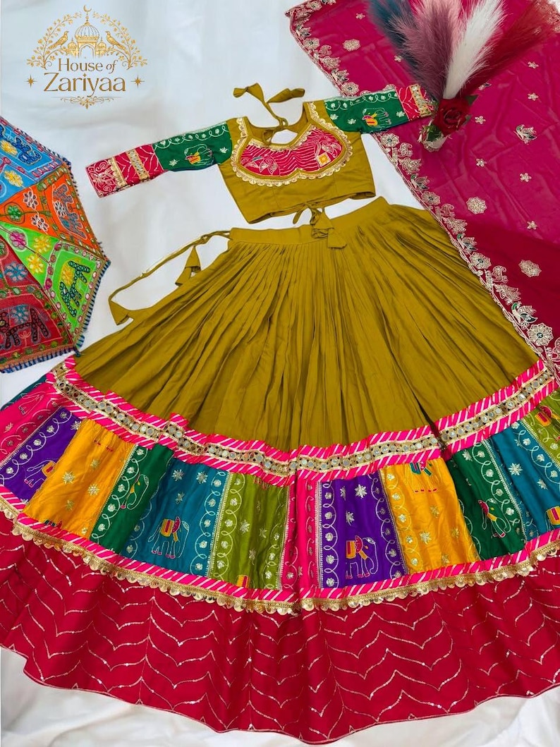 May include: A mustard yellow lehenga choli set featuring a pleated skirt with a multi-colored patchwork border of elephant motifs in green, purple, and yellow, accented with gold trim. The blouse has green sleeves and a pink embroidered neckline.