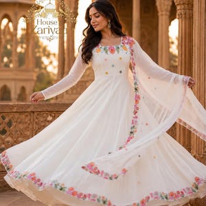 Puede incluir: Un vestido blanco largo hasta el suelo con mangas largas y escote cuadrado, con bordados florales coloridos alrededor del escote y a lo largo del dobladillo. Un dupatta blanco transparente a juego con bordes florales cuelga sobre los brazos.