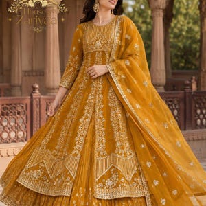 May include: A mustard yellow floor-length gown featuring intricate gold embroidery, a round neckline, and long sleeves. The dress includes a layered skirt with detailed floral patterns and a matching sheer dupatta draped over the shoulder.