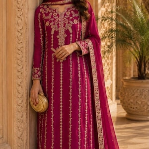May include: A magenta long-sleeved tunic dress featuring intricate gold embroidery in vertical stripes and a detailed floral border at the hem. The garment includes a matching magenta dupatta with a gold embroidered edge and a small gold clutch.