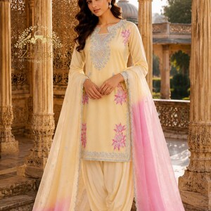 May include: A pale yellow salwar kameez set featuring a long-sleeved tunic with light blue and pink floral embroidery around the neckline and hem. The outfit includes matching loose-fitting trousers and a sheer dupatta with a pink ombre gradient.