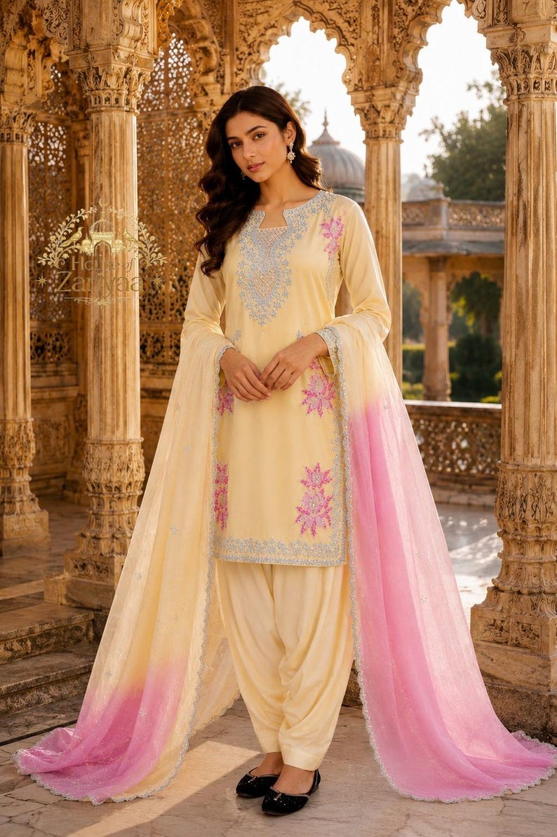 May include: A pale yellow salwar kameez set featuring a long-sleeved tunic with light blue and pink floral embroidery around the neckline and hem. The outfit includes matching loose-fitting trousers and a sheer dupatta with a pink ombre gradient.