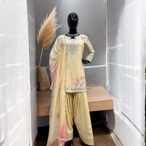 May include: A pale yellow salwar kameez set featuring a long-sleeved tunic with silver embroidery around the neckline and pink floral patterns. The outfit includes matching loose-fitting trousers and a sheer, light yellow dupatta with pink edges.
