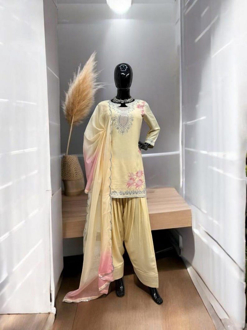 May include: A pale yellow salwar kameez set featuring a long-sleeved tunic with silver embroidery around the neckline and pink floral patterns. The outfit includes matching loose-fitting trousers and a sheer, light yellow dupatta with pink edges.