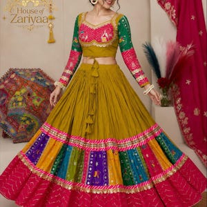 Könnte beinhalten: Ein senfgelbes Lehenga-Choli-Set mit einem bauchfreien Oberteil und langen, grün-rosa gemusterten Ärmeln. Der Rock hat einen breiten, plissierten senfgelben oberen Teil, ein buntes Paneel mit aufwendiger Stickerei und einen dunkelrosa Saum.