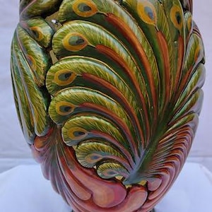 May include: A decorative vase featuring a sculpted peacock feather motif in shades of emerald green, gold, and burnt orange. The exterior surface is textured with layered, overlapping feather shapes that wrap around the entire body of the vessel.
