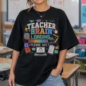 May include: A black t-shirt featuring a colourful graphic with the text "TEACHER BRAIN LOADING... PLEASE WAIT". The design includes a loading bar, a laptop icon, stacks of books, a pencil, and a to-do list, all rendered in vibrant colours.