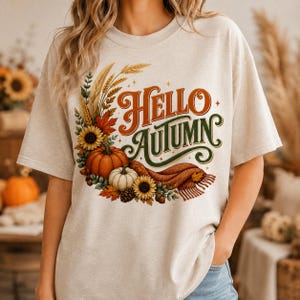 Op de afbeelding: Een beige t-shirt met korte mouwen en een grafische print met de tekst Hello Autumn in oranje en groen schrift. Het ontwerp bevat een halvemaanvormige opstelling van tarwe, zonnebloemen, oranje en witte pompoenen en een bruine sjaal.
