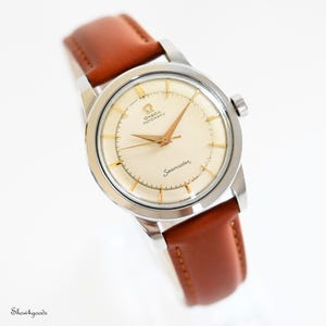 Può includere: Un orologio da polso automatico Omega Seamaster con cassa rotonda in acciaio inossidabile color argento, quadrante color crema, indici e lancette color oro e un cinturino in pelle marrone liscia con cuciture visibili sui bordi.