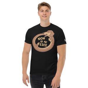Può includere: Una t-shirt nera a maniche corte con la grafica di un bassotto marrone arrotolato in cerchio che si morde la coda. All'interno del cerchio, il testo "NON SE NE ESCE" è scritto in lettere maiuscole giallo chiaro. Il disegno è centrato sul petto.