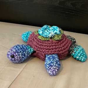 May include: A handmade crochet turtle plush featuring a dark purple shell topped with a blue and green floral accent. The head and four flippers are crafted from variegated yarn in shades of blue, purple, and white, with small black bead eyes.