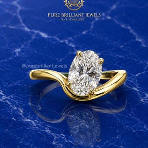 May include: A gold ring featuring a single oval-cut clear gemstone held by four prongs on a smooth, curved band. The text PURE BRILLIANT JEWELS FINE JEWELLERY and @purebrilliantjewels are visible above the ring on a blue marble-patterned surface.