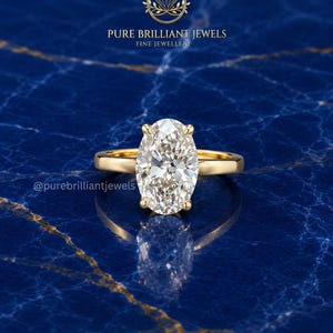 Può includere: Un anello in oro con una grande pietra preziosa trasparente a taglio ovale fissata da quattro griffe. Il testo PURE BRILLIANT JEWELS FINE JEWELLERY e @purebrilliantjewels è visibile. L'anello presenta una fascia in oro giallo lucido.