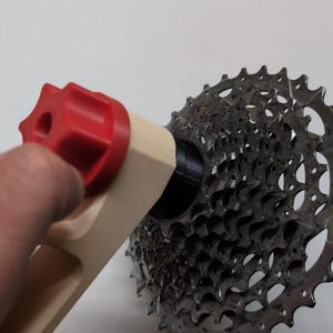 Könnte beinhalten: Ein Reinigungswerkzeug für Fahrradkassetten mit einem beigen Griff, einem roten Einstellknopf und einem schwarzen zylindrischen Befestigungspunkt. Das Werkzeug ist an einer silberfarbenen Metall-Fahrradkassette mit mehreren Zahnrädern befestigt.
