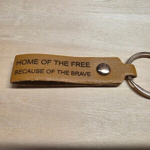 May include: A tan leather keychain featuring a silver metal ring and a silver rivet. The leather strap is engraved with the text HOME OF THE FREE BECAUSE OF THE BRAVE in a dark, sans-serif font. The keychain is rectangular with a rounded end.