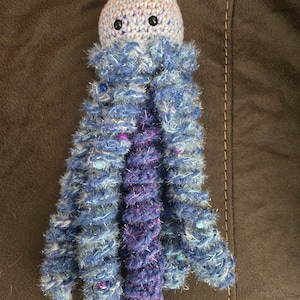May include: A crocheted jellyfish toy with a round, light-colored head featuring two black bead eyes. The body has multiple long, spiraling tentacles made of blue and purple fuzzy yarn with metallic silver strands and small embedded beads.