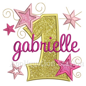 May include: Gold glitter number one with pink glitter stars and the name gabrielle in pink cursive lettering.