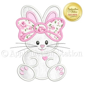 May include: A white bunny with pink ears and a pink bow with a floral pattern. The bunny is sitting with its paws in front of it. The bunny has a small pink heart on its chest. The image is a digital embroidery design.