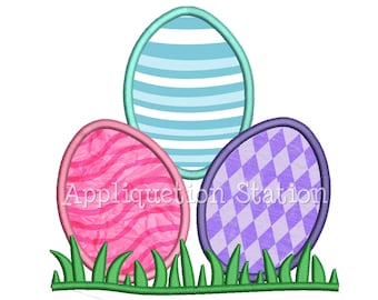 May include: Three Easter eggs in a row, with a blue and white striped egg in the middle, a pink and white striped egg on the left, and a purple and white checkered egg on the right. The eggs are sitting on a bed of green grass.