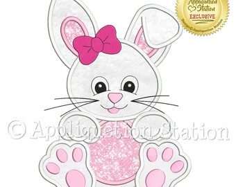 May include: White bunny applique with pink ears, a pink bow, and pink paws. The bunny is sitting with a pink glitter circle behind it.