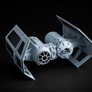 Puede incluir: Un modelo de plástico gris claro de una nave espacial TIE fighter que presenta una cabina esférica central con una ventana circular, flanqueada por dos grandes alas rectangulares planas con un patrón de rejilla y detalles mecánicos.
