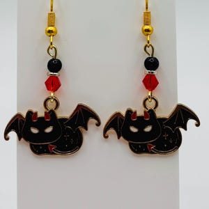 May include: A pair of dangle earrings featuring gold-toned hooks, a black bead, a silver spacer, and a faceted red crystal bead. The charms are black enamel bat-like creatures with red horns, red tails, and white eye outlines, suspended from the beads.