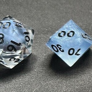 May include: A set of seven polyhedral dice featuring a translucent light blue and white marbled resin finish with embedded glitter particles. Each die has clear, sharp edges and bold black numerals engraved on the faces for high visibility.