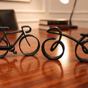 Könnte beinhalten: Zwei schwarze, minimalistische Fahrradskulpturen in einem durchgehenden Liniendesign. Jedes Stück verfügt über kreisförmige Räder, einen Rahmen, einen Sattel und einen Lenker und ist als dekoratives Tischornament mit matter Oberfläche gestaltet.